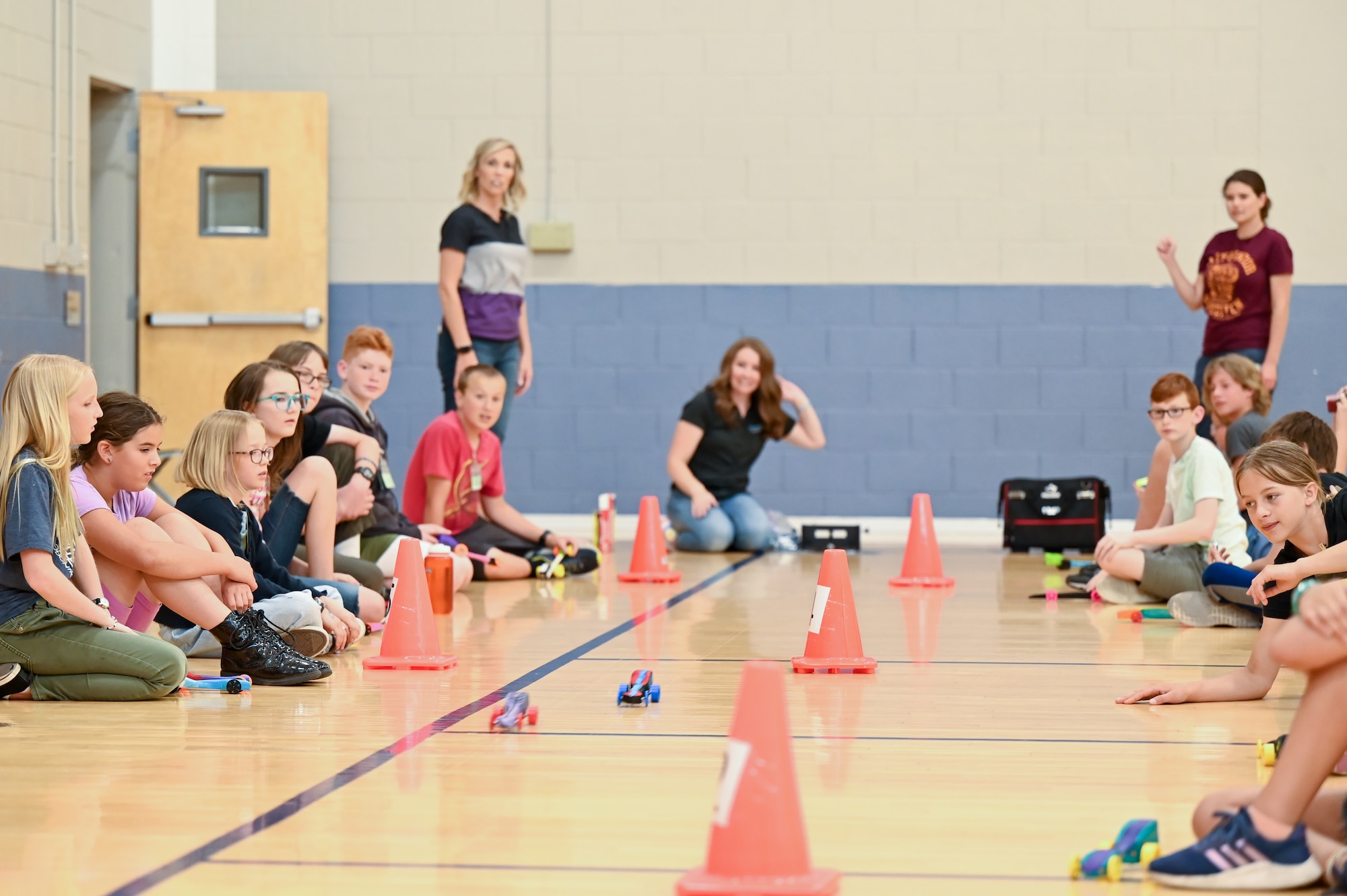 Two CO2 dragsters race on the gym floor between orange cones flanked on both sides by students watching on.