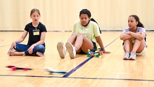 Three students sit on the gym floor as two CO2 dragsters whiz by in front of them.