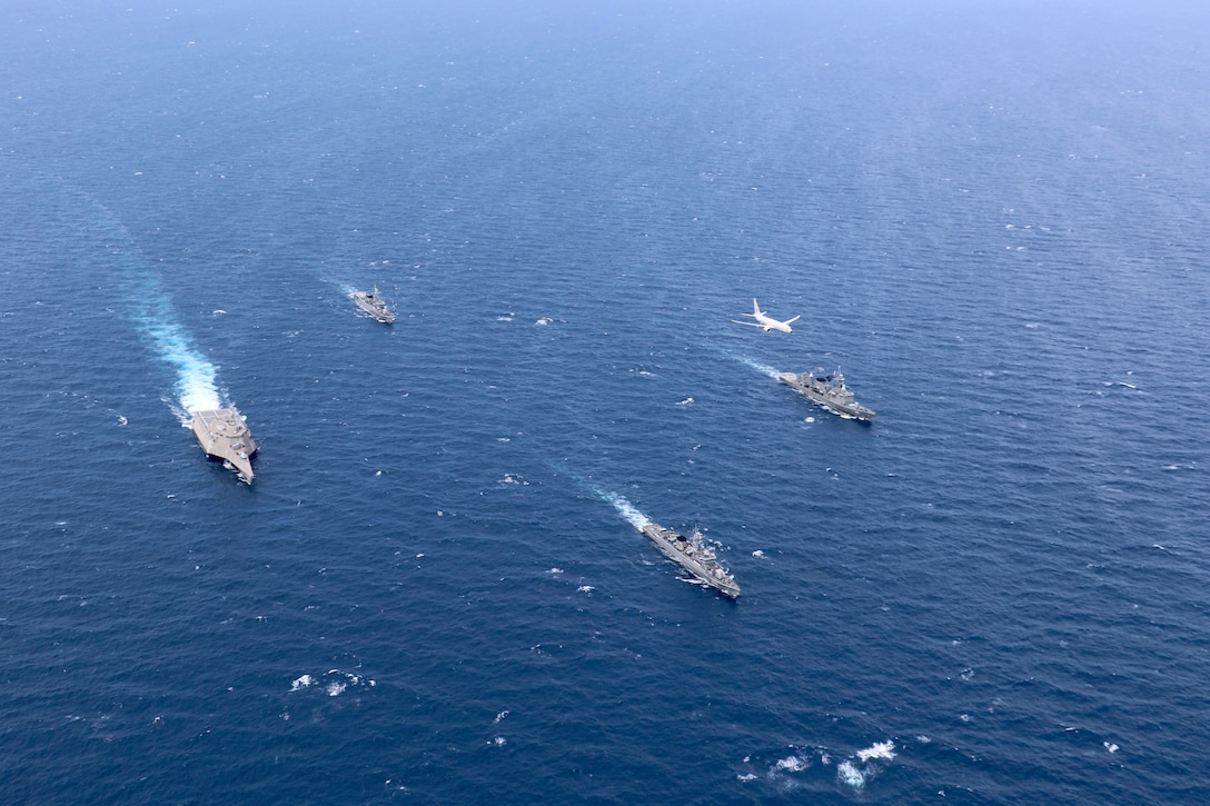 Ships sail in formation as an aircraft flies above.