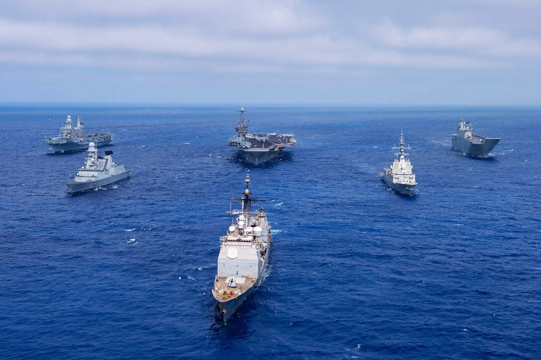 Five naval ships sail in a wedge formation ahead of an aircraft carrier.