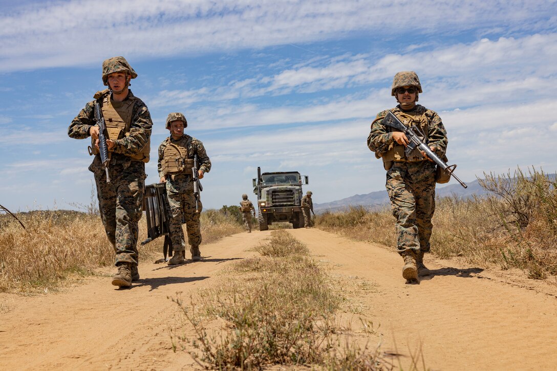 Three Marines with weapons walk down a dirt road with a truck behind them.