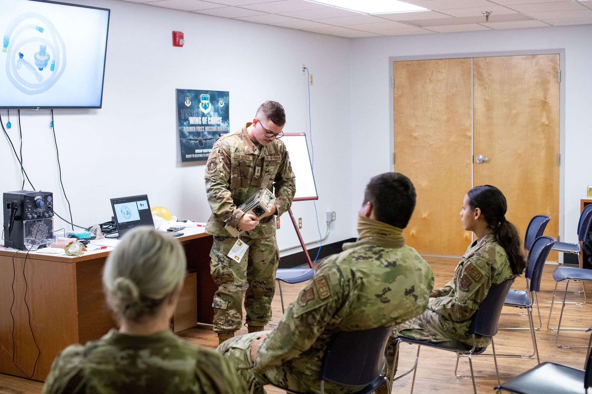 Hathorn, standing, gestures to a ventilator machine he's holding while Airmen sitting in front of him look on