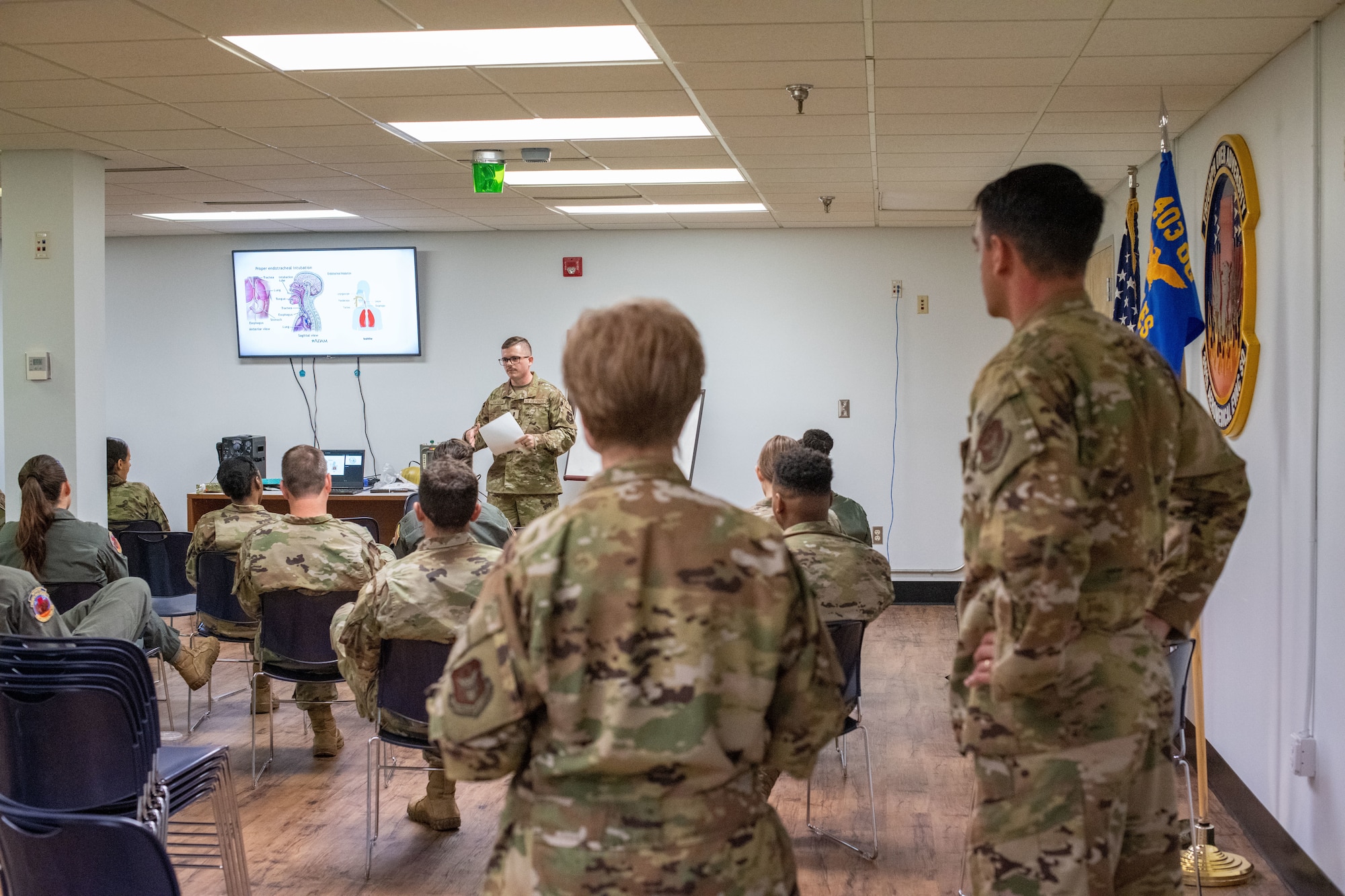 Schreffler and Rubio stand in the foreground facing forward as a class is seated listening to the instructor at the front of the room