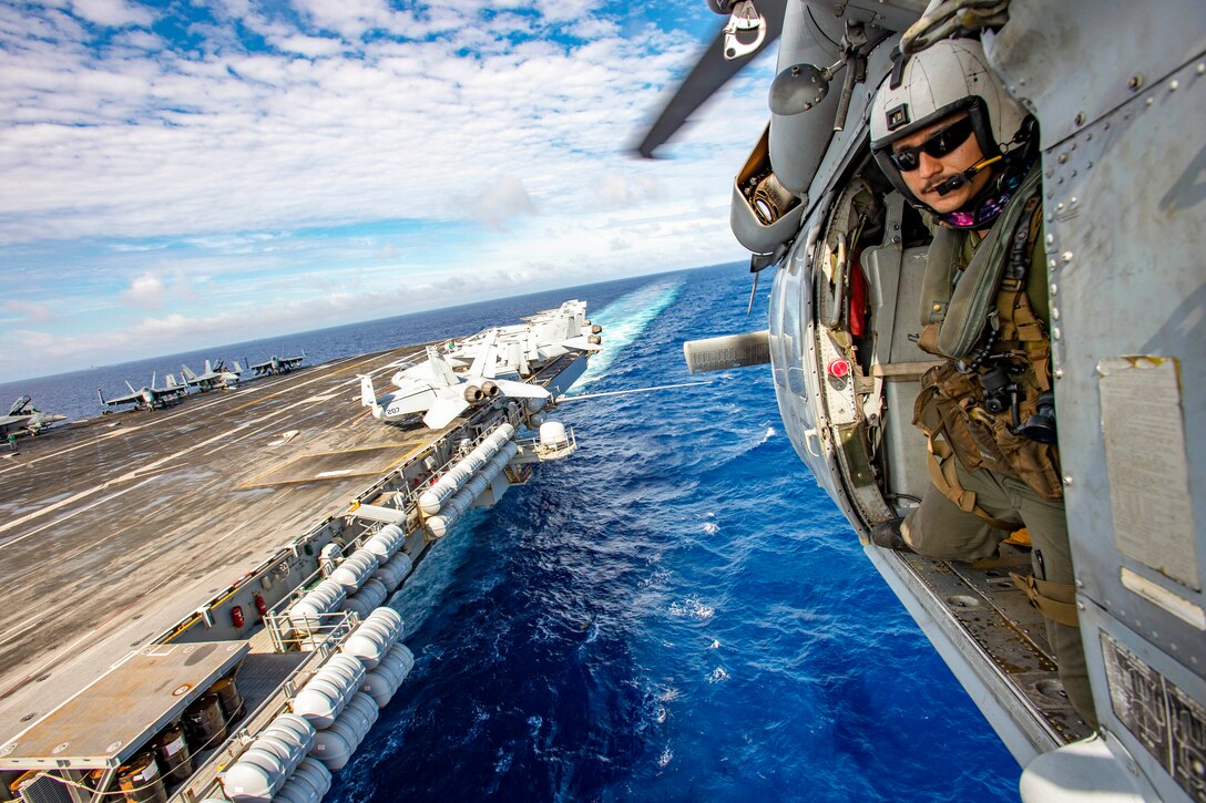 A sailor stands in the doorway of a helicopter flying next to a ship at sea.