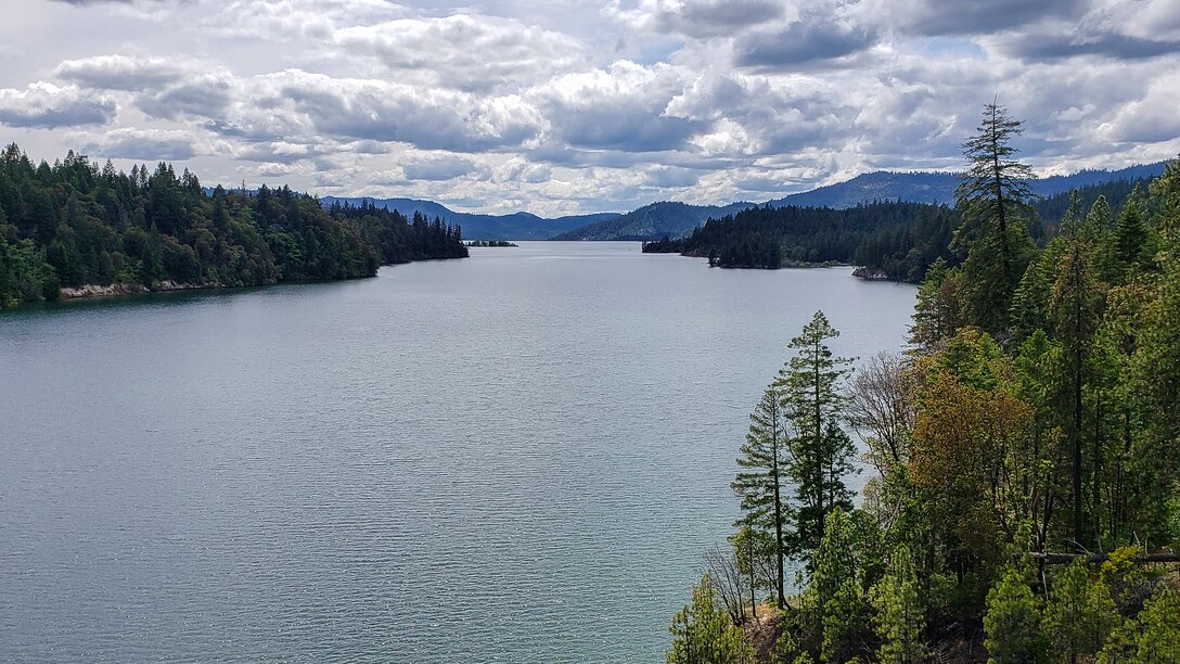 Gray and white clouds loom over a calm lake surrounded by trees and hills on an overcast day.