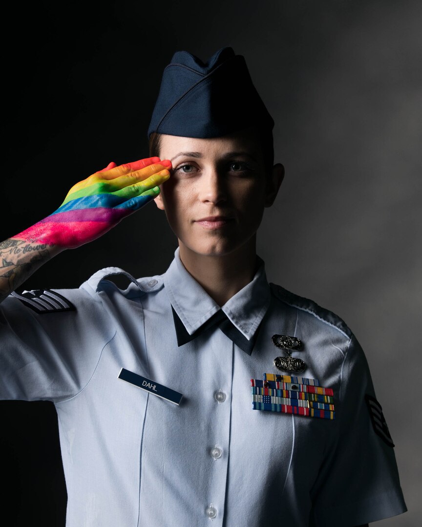 A service member poses for a portrait and salutes with a rainbow-painted hand.