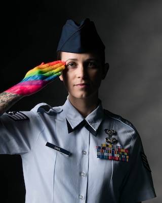 A service member poses for a portrait and salutes with a rainbow-painted hand.