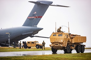 Service members operate and stand near two military vehicles. A large airplane in the background has an open cargo hold.