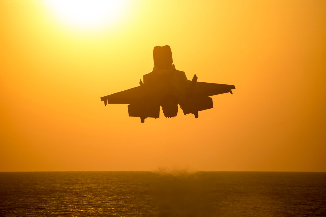 An aircraft launches over a body of water under a sunlit sky.