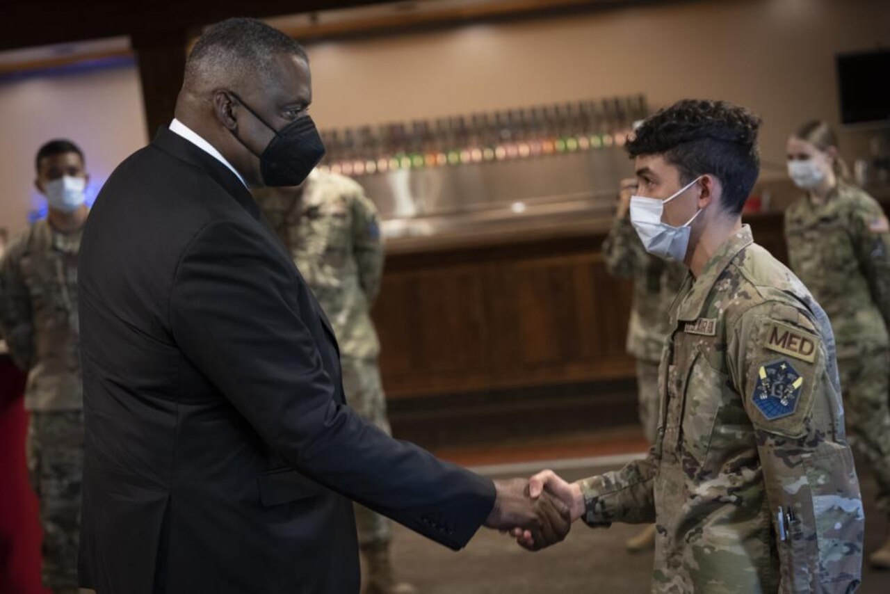 A man in a business suit wearing a face mask shakes hands with a uniformed service member, who is also wearing a face mask.