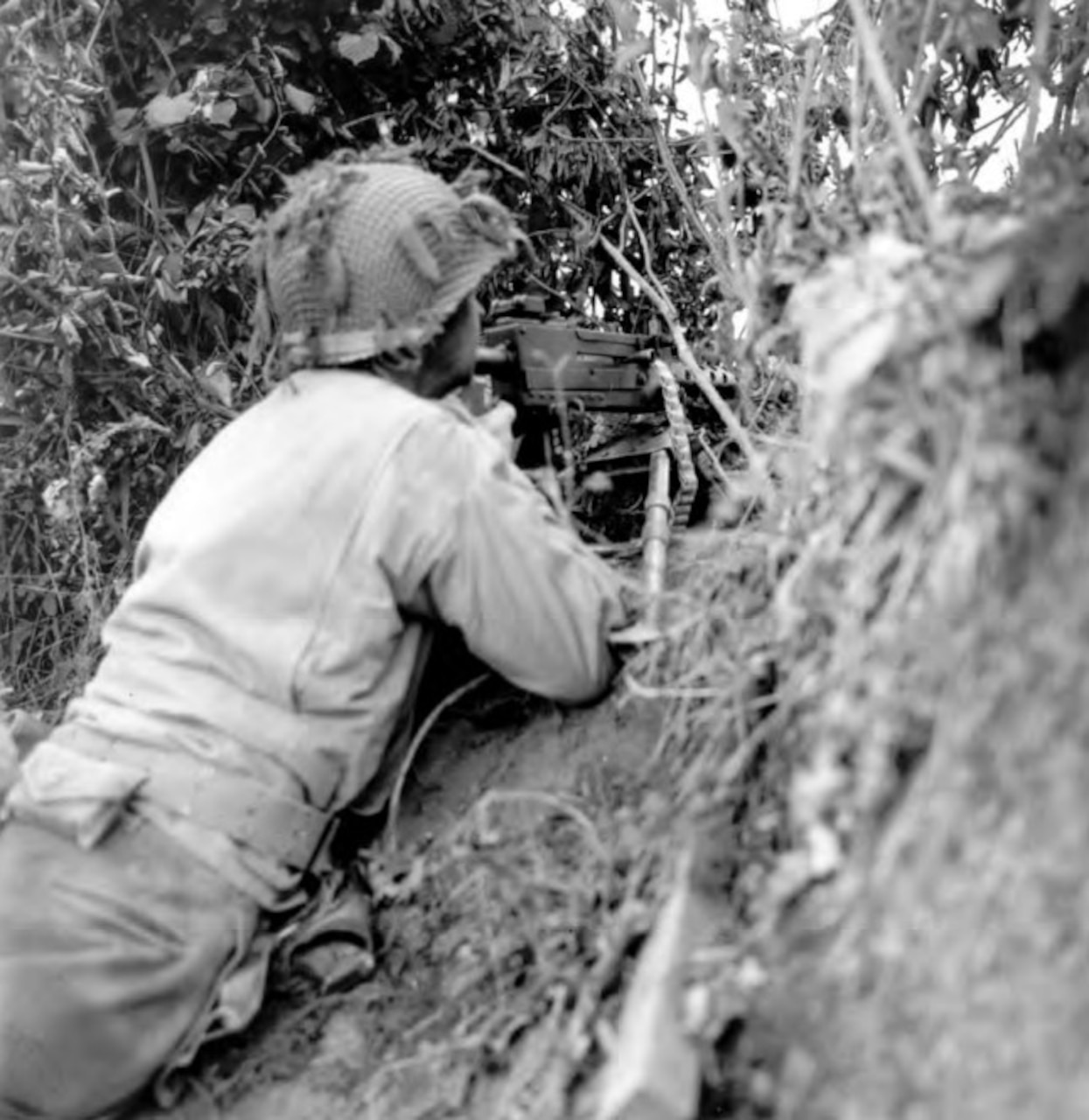 A soldier with a machine gun points it over a hedgerow.