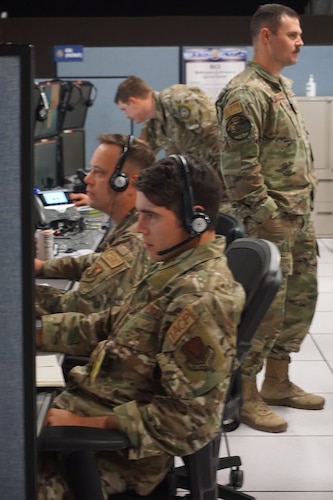 photo of two USAF Airmen wearing headsets working at computers, two USAF Airmen standing