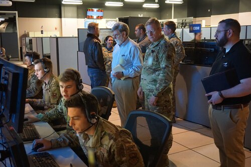 photo of four USAF Airmen working at computers with three USAF Airmen standing behind them watching, in the background four additional USAF Airmen are talking in a group
