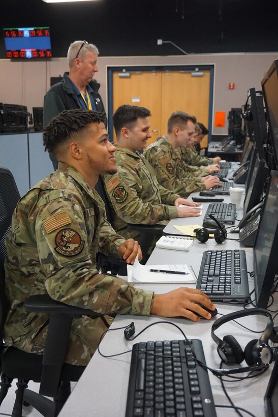 photo of four USAF sitting working on computers with one USAF Airmen standing behind them observing