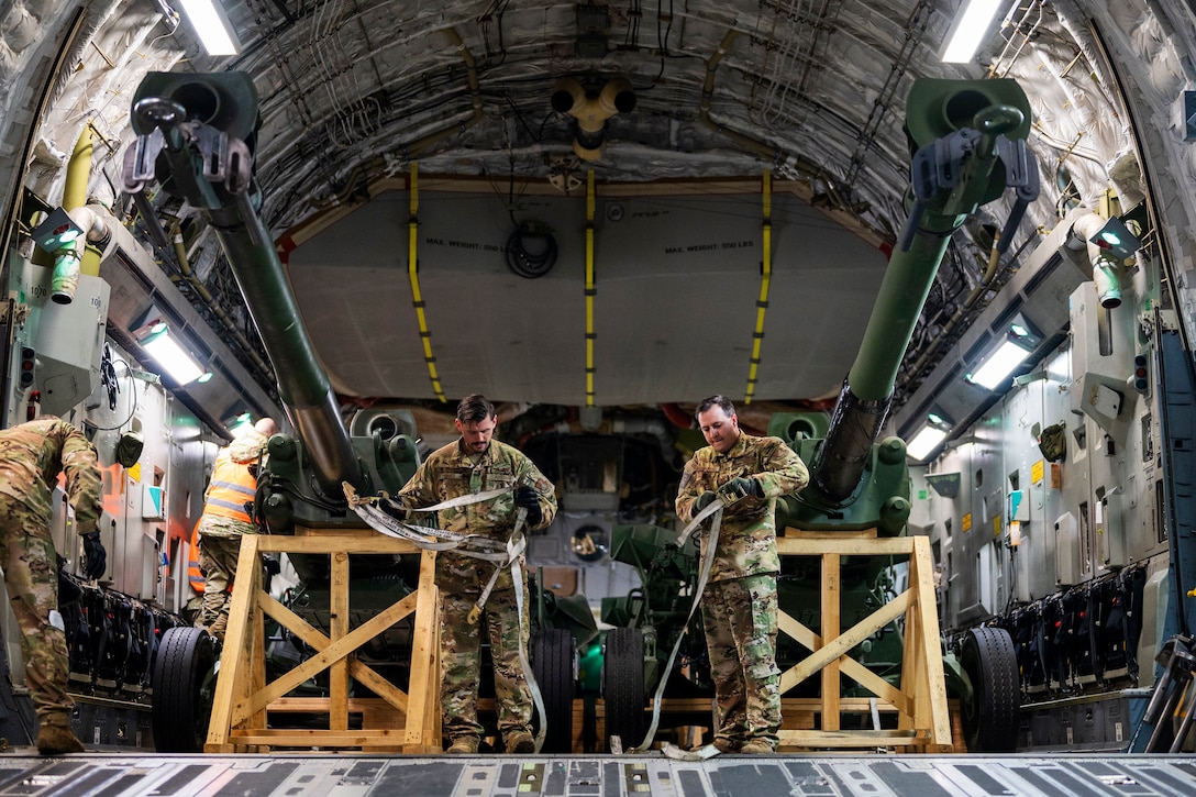Two service members remove straps from two large pieces of artillery in  the cargo holding area of an airplane.