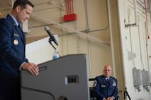 Chief Master Sgt. Peter Webb sits on stage while Col. Kevin Merrill stands at podium during a retirement ceremony.