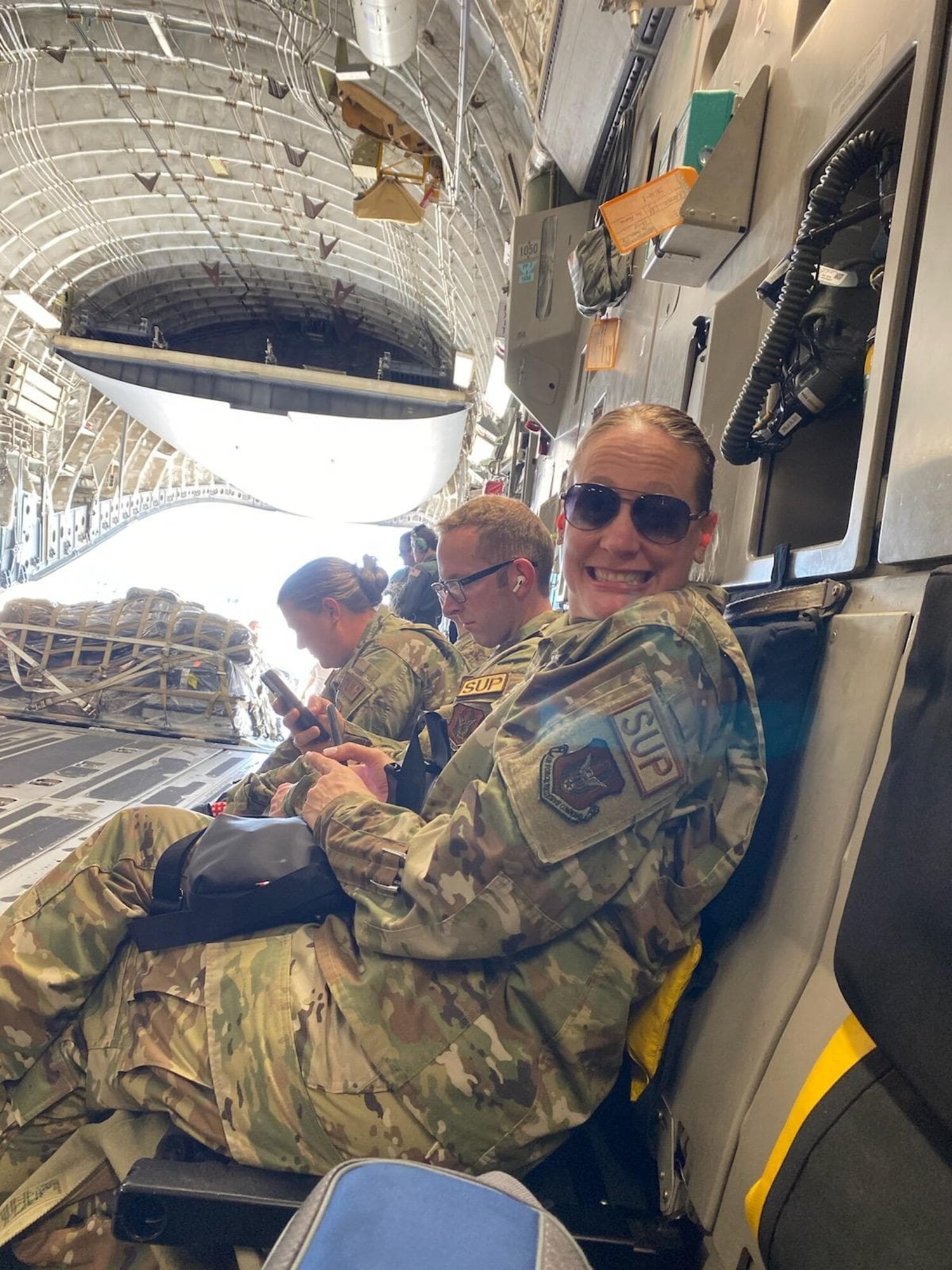 A woman in camouflage and aviator sunglasses smiles at the camera in the back of a cargo aircraft.
