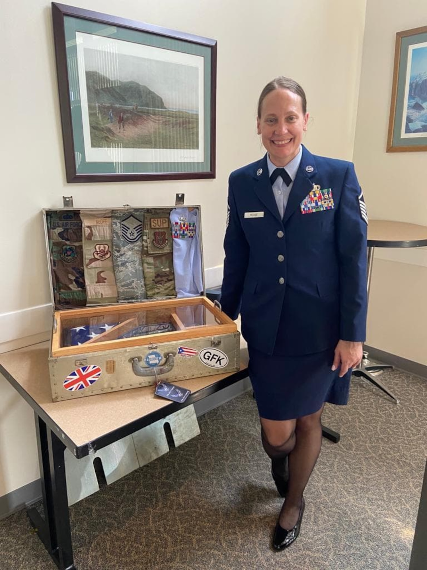 A woman Air force service dress stands next to a suitcase on a table. The suitcase contains five different Air Force uniforms, representing the uniform styles worn over the past two decades, and a folded American flag.
