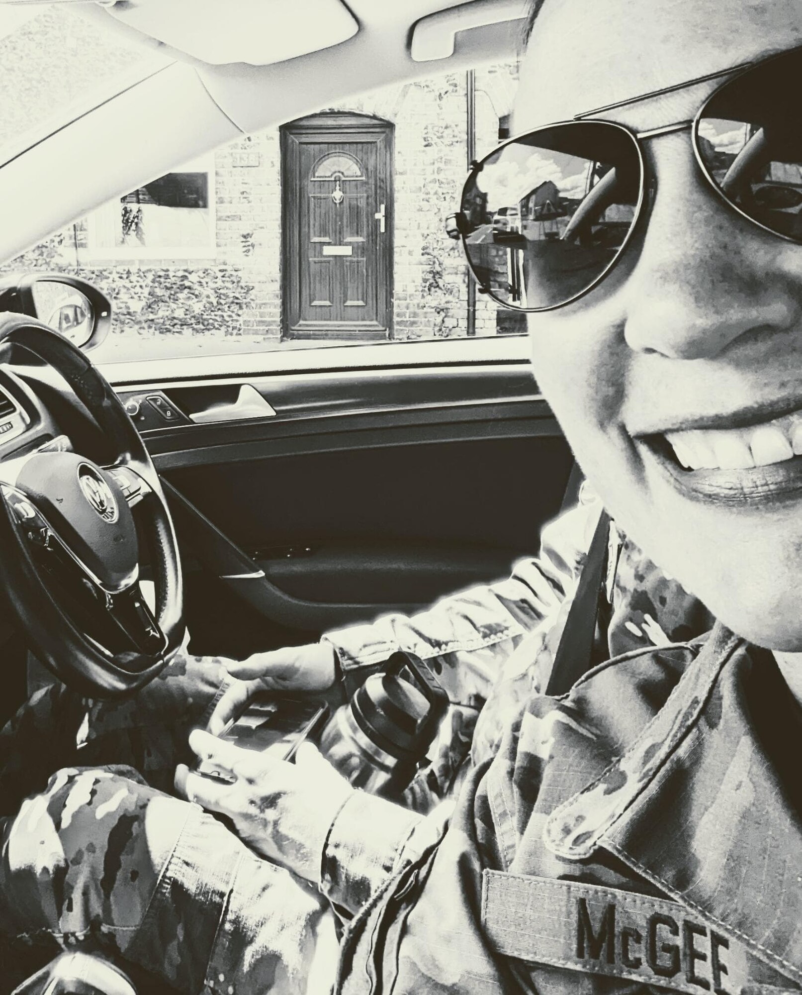 A black-and-white photo of a smiling woman in aviator sunglasses in the passenger seat of a car. The front door of a house can be seen through the driver's window.