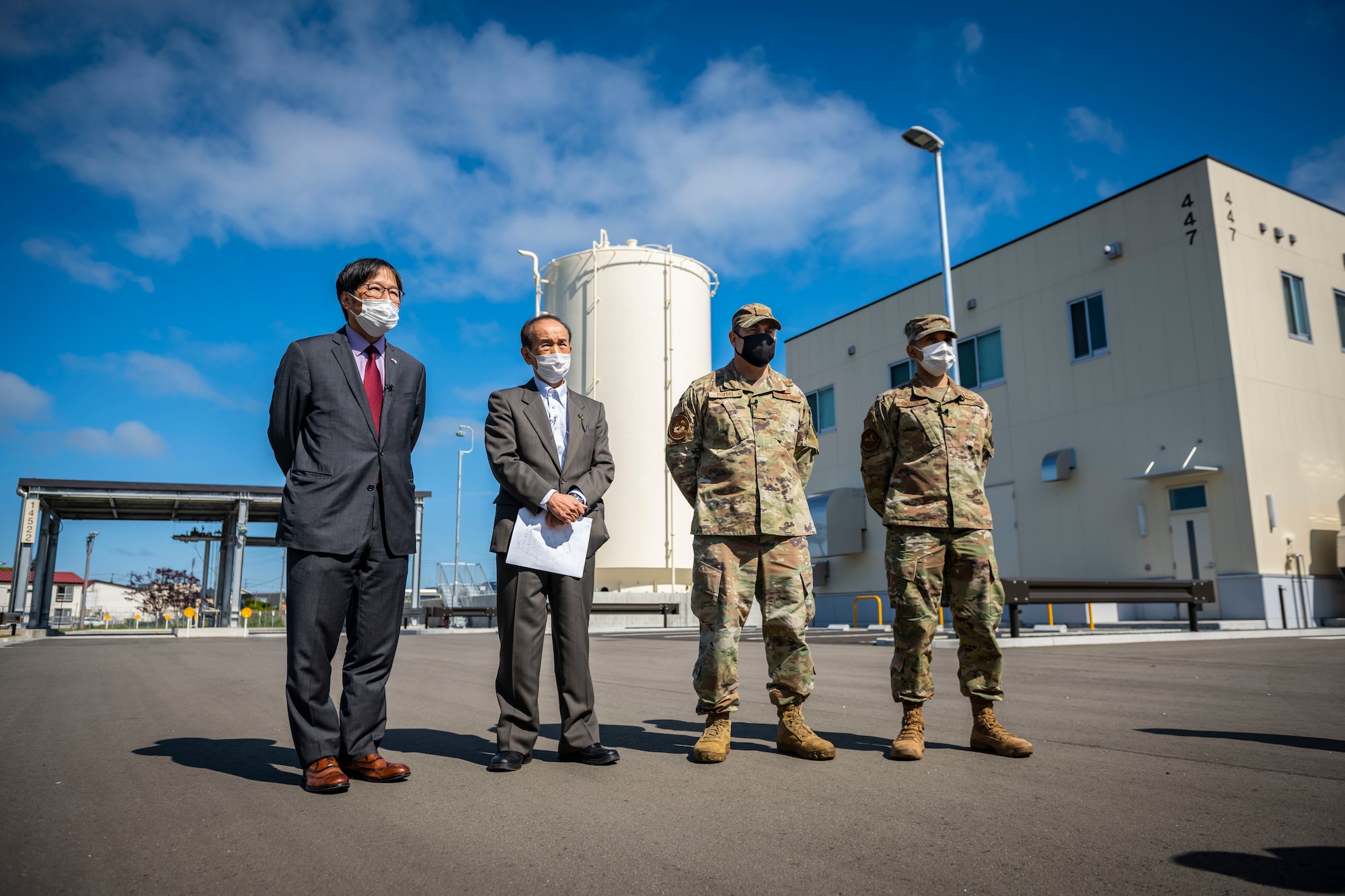 military members in uniform and members in suits stand in front of a gas plant.