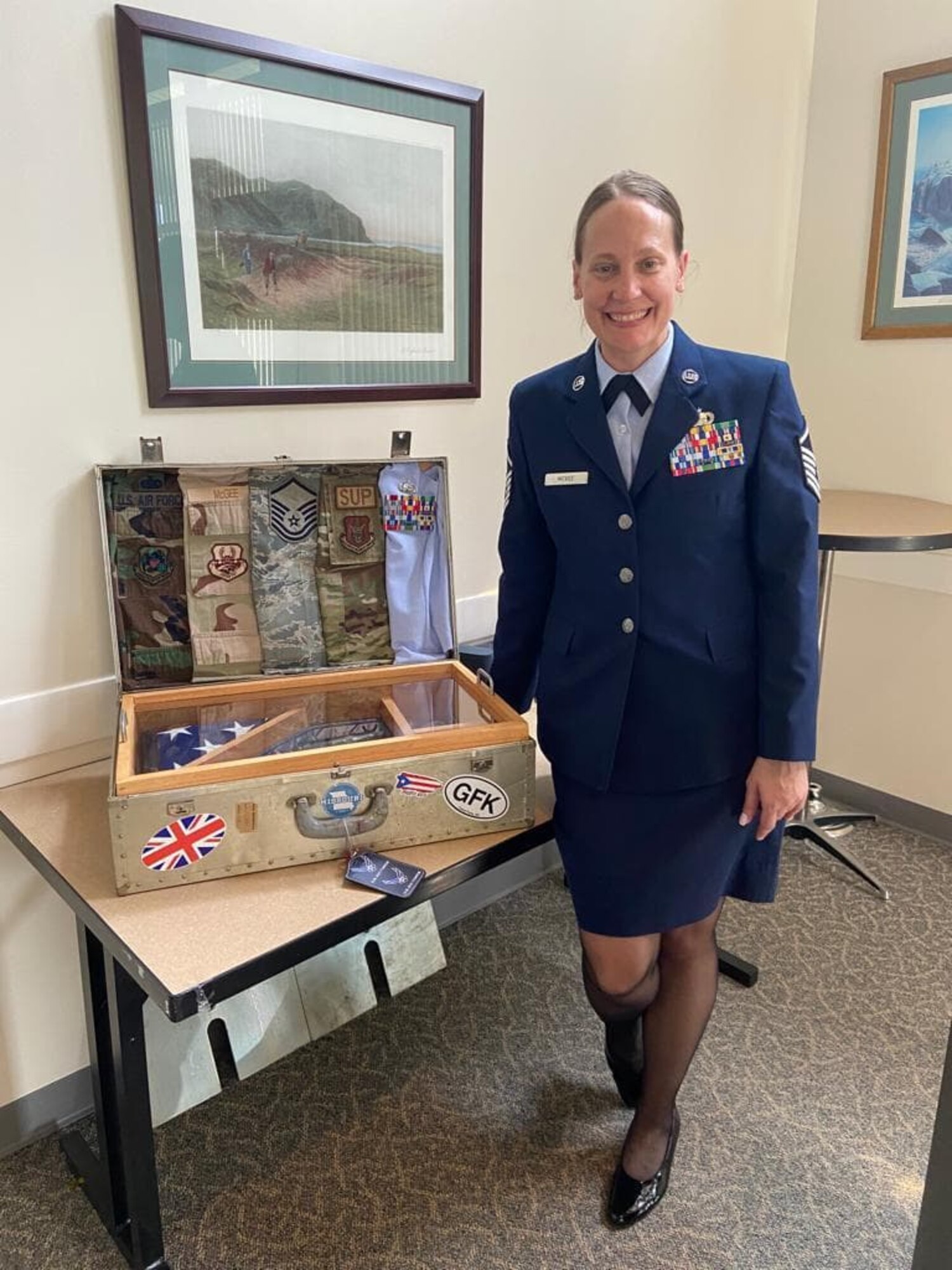 A woman Air force service dress stands next to a suitcase on a table. The suitcase contains five different Air Force uniforms, representing the uniform styles worn over the past two decades, and a folded American flag.