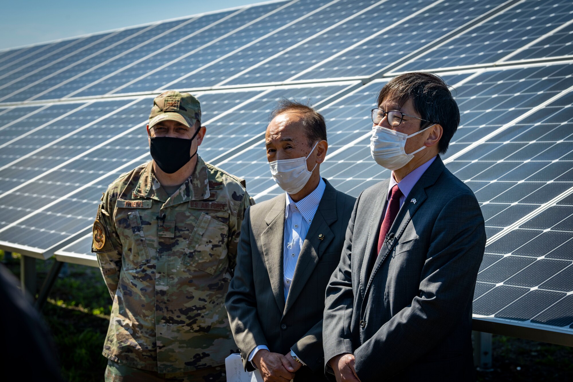 Military member in uniform and members in suits stand in front of a solar panel.