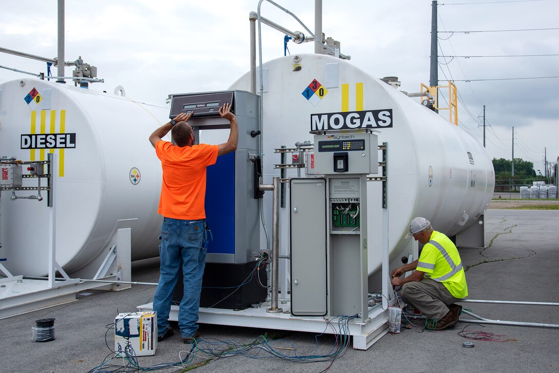Two men in bright OSHA safety gear work near three large white cylindrical fuel tanks.