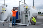 Two men in bright OSHA safety gear work near three large white cylindrical fuel tanks.