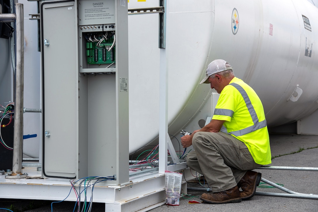 A man in bright OSHA safety gear and a hardhat works near a large white cylindrical fuel tank.