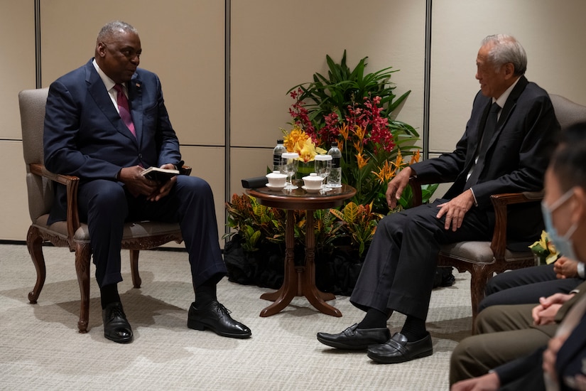 Two seated men wearing suits talk to each other.