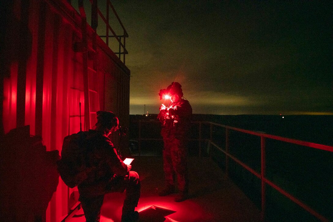 A U.S. airman monitors a screen while an Estonian kneels to look at an object illuminated by red light.