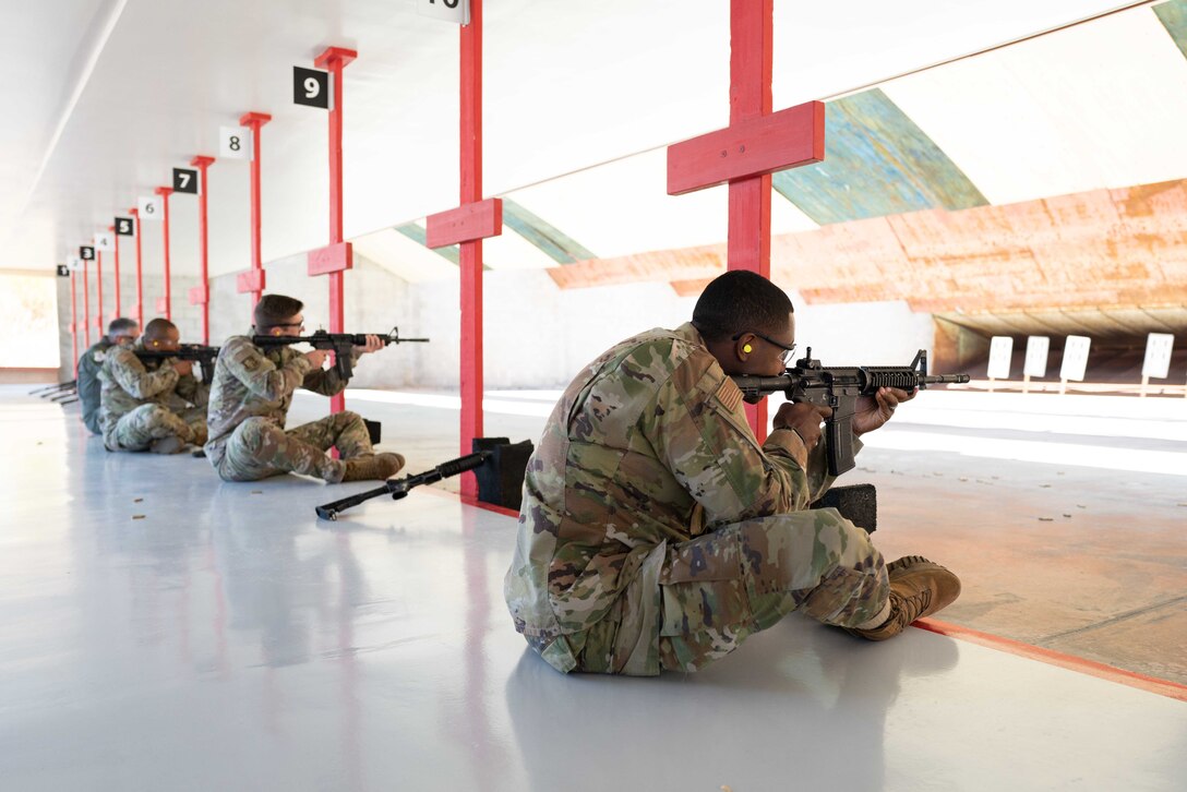 Airmen sit in a line while firing rifles at a range.