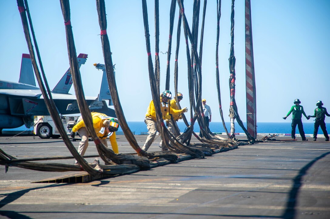A group of sailors work on vertical cables on the flight deck of a ship.
