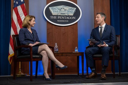 A woman and a man sit facing each other. A U.S. flag and a sign that says "The Pentagon -- Washington" are in the background.