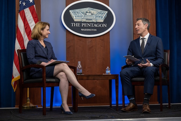 A woman and a man sit facing each other. A U.S. flag and a sign that says "The Pentagon -- Washington" are in the background.