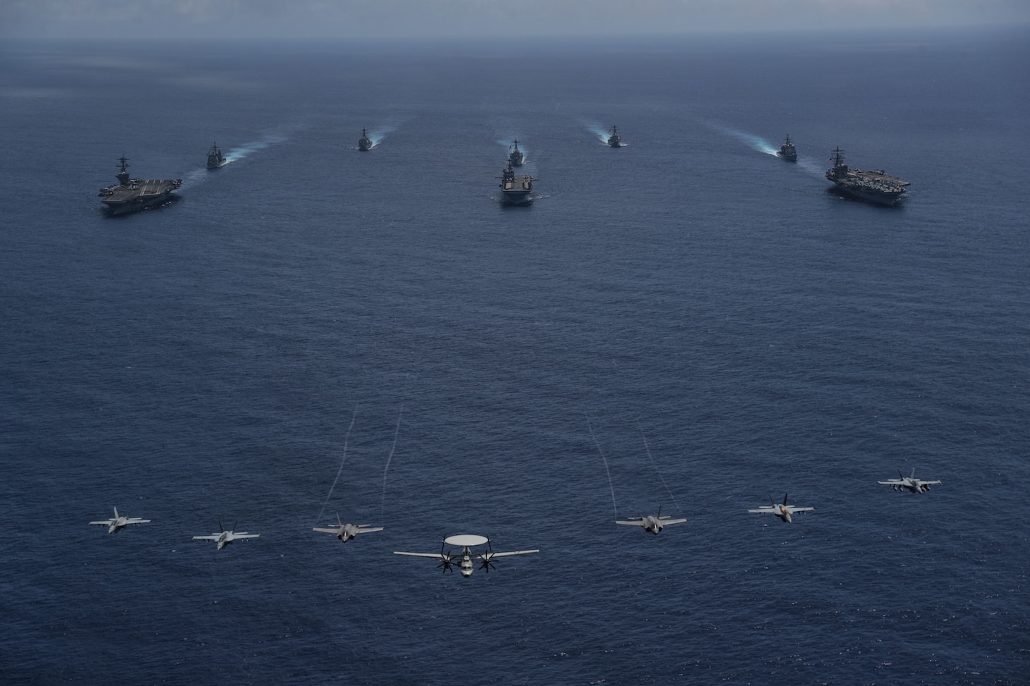 PHILIPPINE SEA (June 12, 2022) Aircraft from Carrier Air Wing (CVW) 9 fly over the Nimitz-class aircraft carrier Abraham Lincoln (CVN 72), front left, America-class amphibious assault ship USS Tripoli (LHA 7), front center, Nimitz-class aircraft carrier USS Ronald Reagan (CVN 76), front right, Ticonderoga-class guided-missile cruiser USS Mobile Bay (CG 53), middle left, Arleigh Burke-class guided-missile destroyer USS Benfold (DDG 65), middle center, Ticonderoga-class guided-missile cruiser USS Antietam (CG 54), middle right, Arleigh Burke-class guided-missile destroyer USS Spruance (DDG 111), back left, and Arleigh Burke-class guided-missile destroyer USS Fitzgerald (DDG 62), back right, as they sail in formation during Valiant Shield 2022 (VS22). VS22 is a U.S.-only, biennial field training exercise (FTX) focused on integration of joint training in a multi-domain environment. This training builds real-world proficiency in sustaining joint forces through detecting, locating, tracking, and engaging units at sea, in the air, on land, and in cyberspace in response to a range of mission areas. (U.S. Navy photo by Mass Communication Specialist 3rd Class Thaddeus Berry)