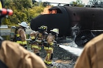 Three 316th Civil Engineer Squadron firefighters pull a gushing fire hose toward a simulated aircraft fire during interagency training at Joint Base Andrews, Md., May 2, 2022. The team trained in a live-burn exercise to practice firefighting techniques and procedures in a realistic environment.