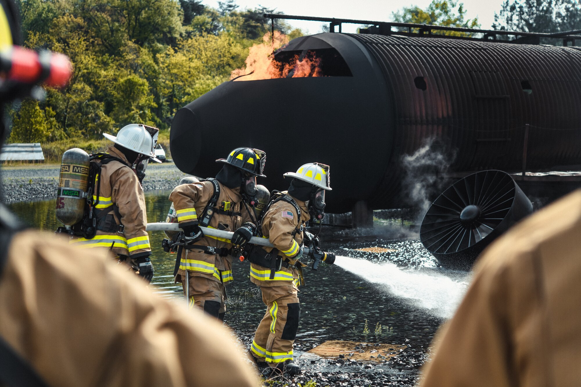 Three 316th Civil Engineer Squadron firefighters pull a gushing fire hose toward a simulated aircraft fire during interagency training at Joint Base Andrews, Md., May 2, 2022. The team trained in a live-burn exercise to practice firefighting techniques and procedures in a realistic environment.