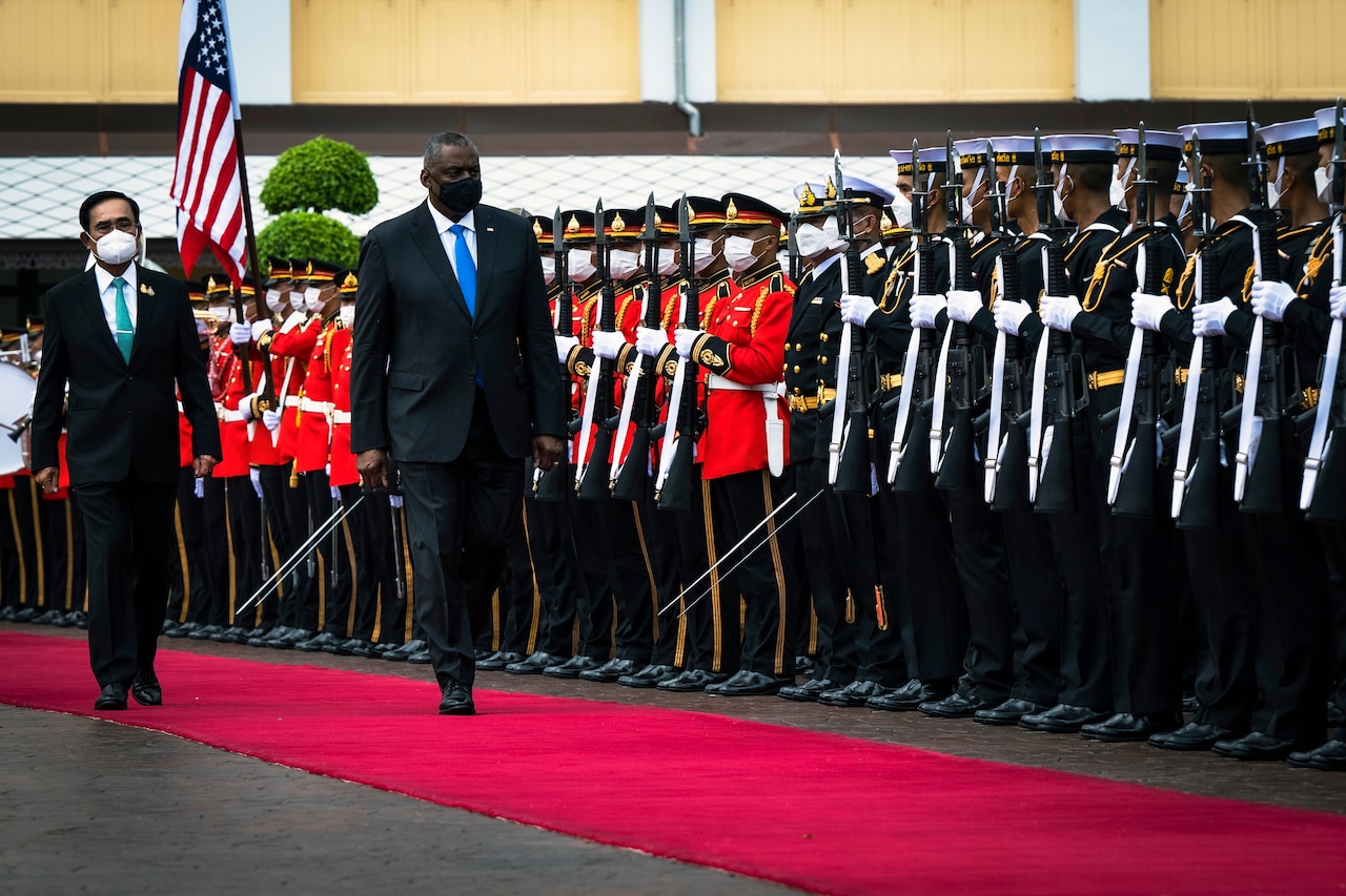 Two men in business attire walk down a red carpet alongside a formation of service members in military formal attire.