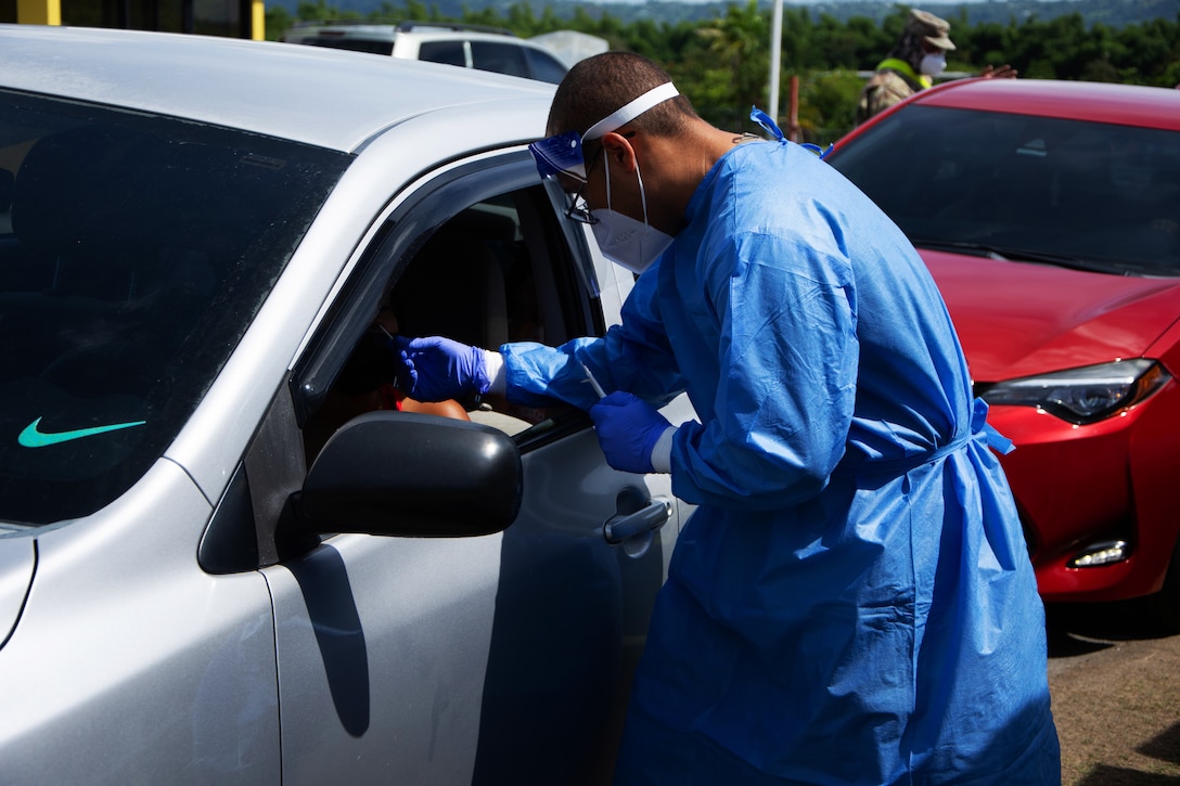 A man wearing medical personally protective equipment holds a nasal swab while leaning into a vehicle.