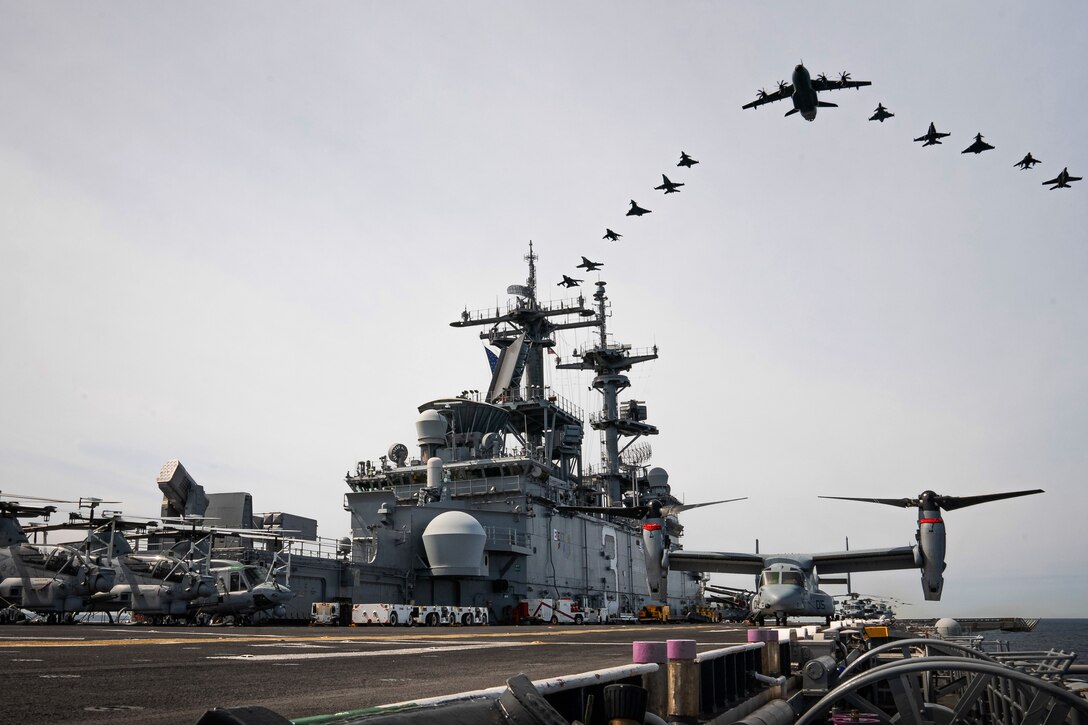 Aircraft fly in a V formation over a naval ship.