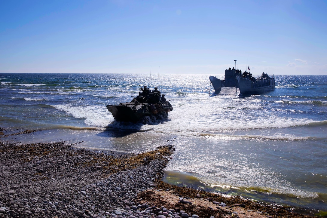 A light armed vehicle disembarks a landing craft in a sea and drives onto shore.