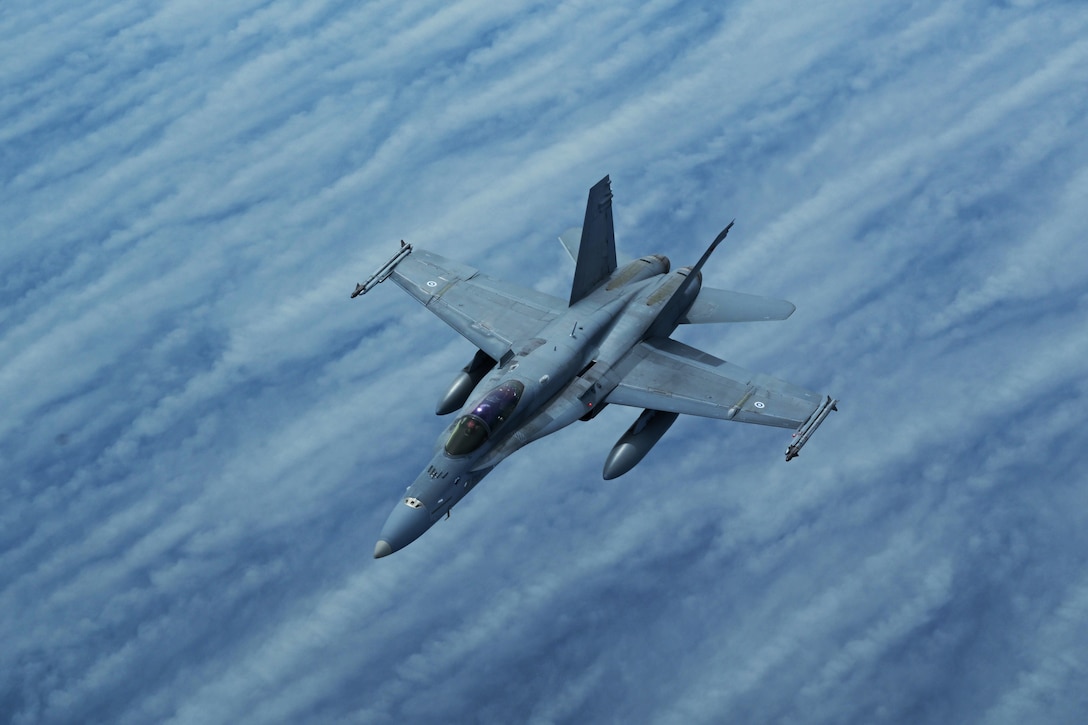 A Navy Superhornet aircraft flies in a blue sky over clouds.