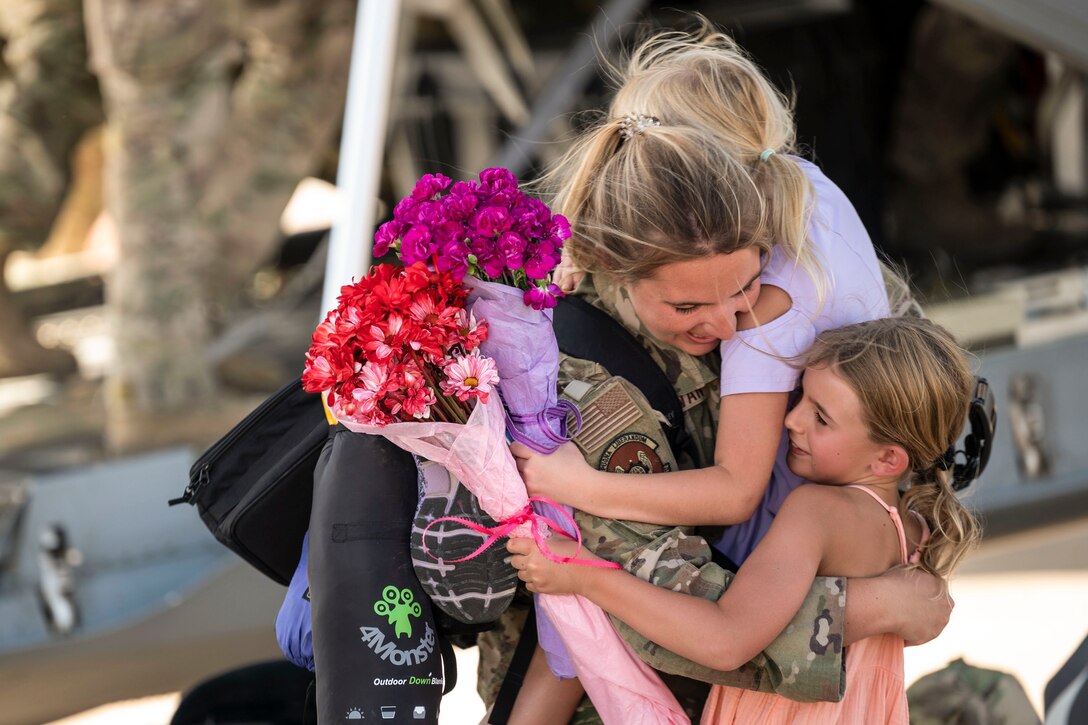 Two children hug an airman while holding flowers in their hands.