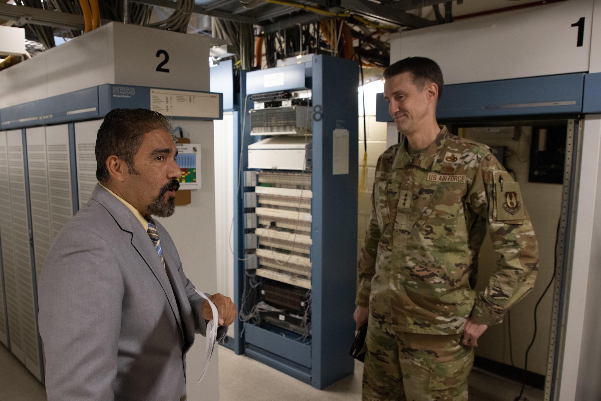 Man in suit talking to Airman in front of phone equipment