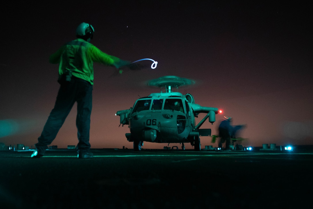A sailor waves a light wand in front of a helicopter at night.
