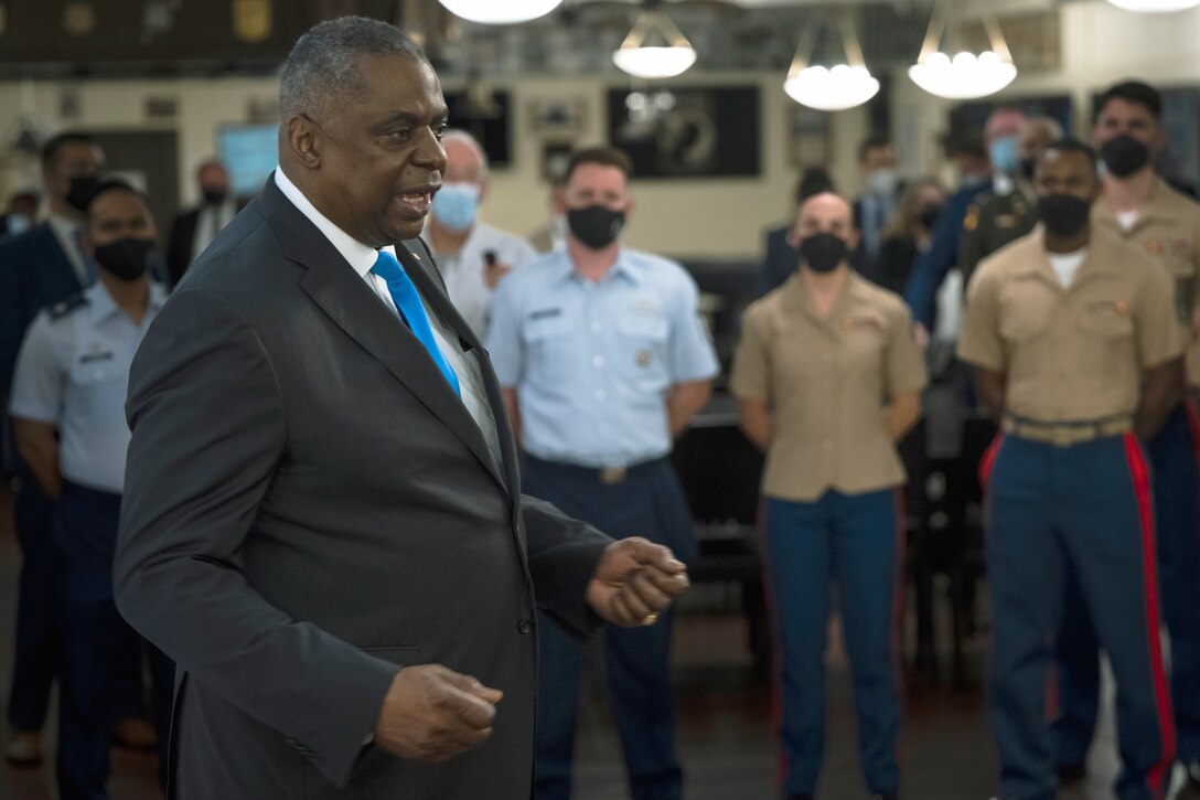 A man in business attire speaks in front of a group of uniformed service members standing at parade rest.