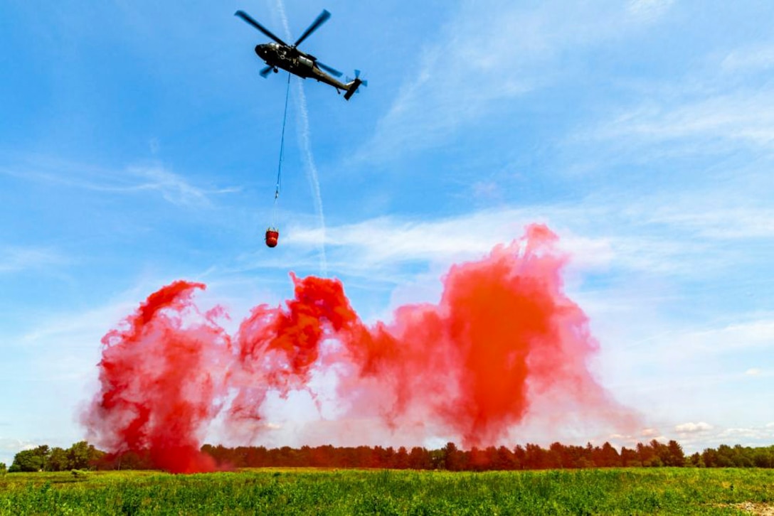 A helicopter carrying a bucket flies over red smoke.