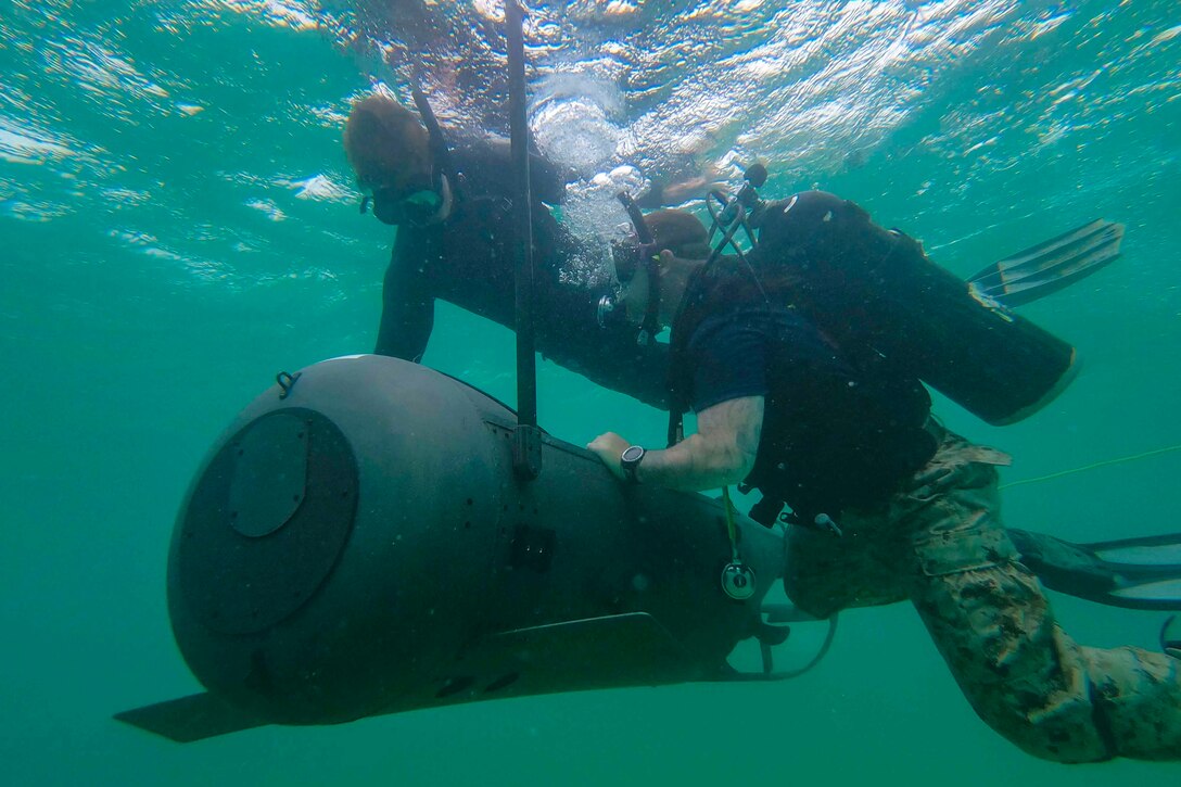 Sailors dive underwater with a piece of equipment.