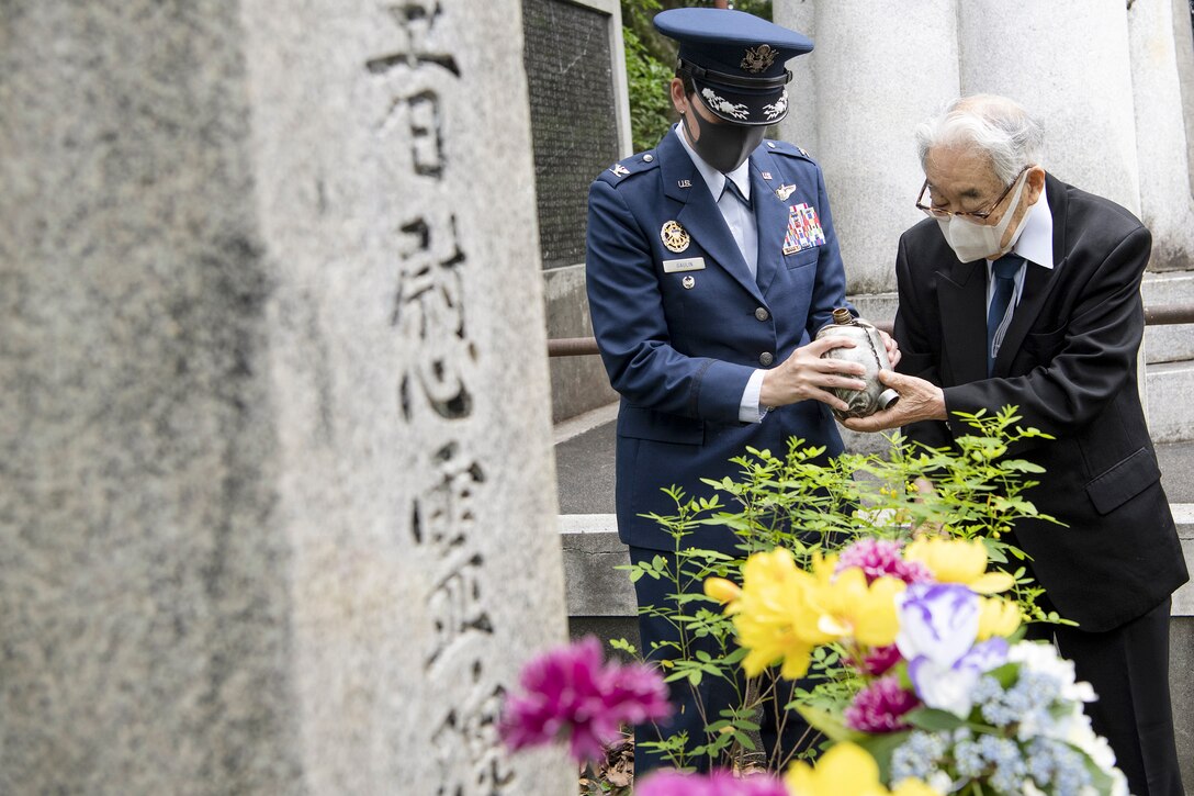 An airman and a civilian hold a canteen at a monument adorned with flowers.